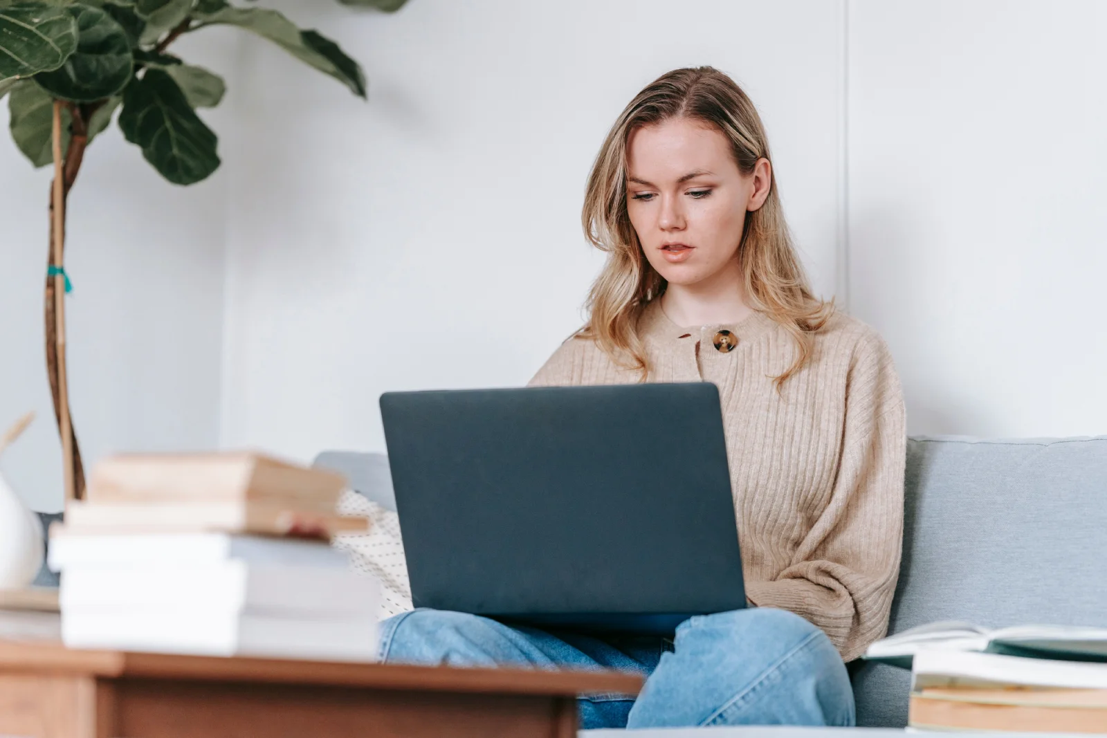 Focused young woman using laptop for study at home, surrounded by books and cozy decor.