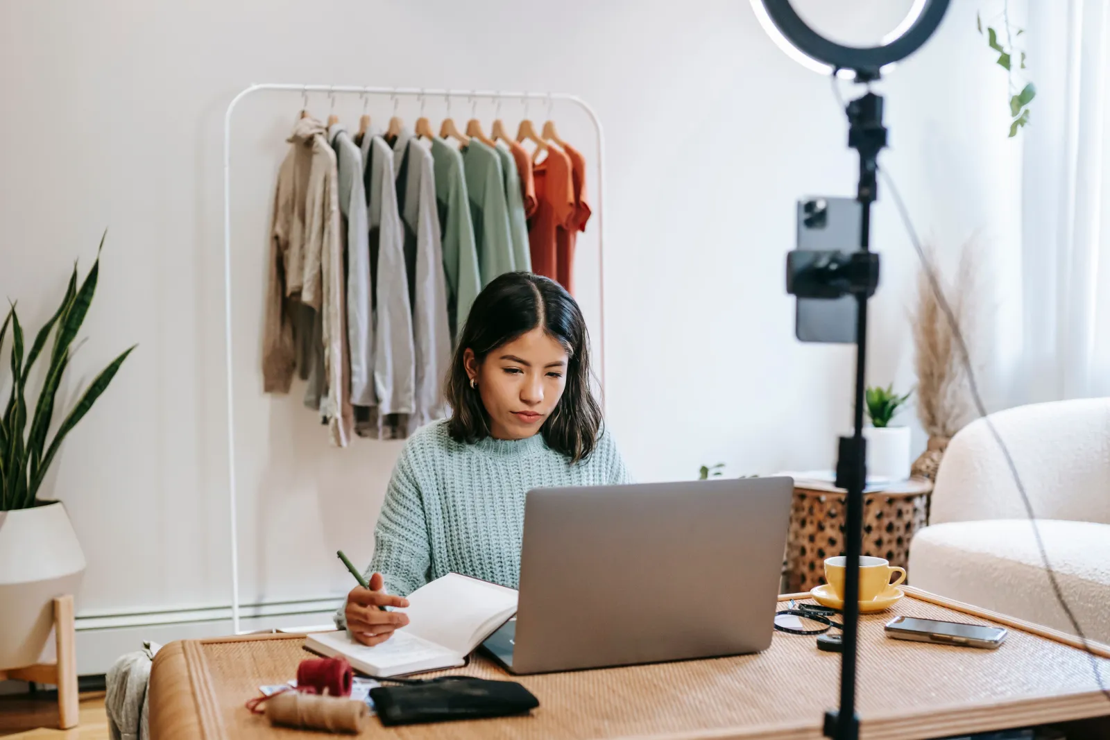 Focused young woman working on laptop in home office, surrounded by clothing rack and camera setup