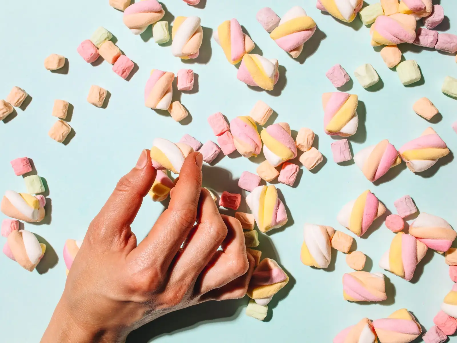 A hand reaches for colorful marshmallows scattered on a light blue surface.