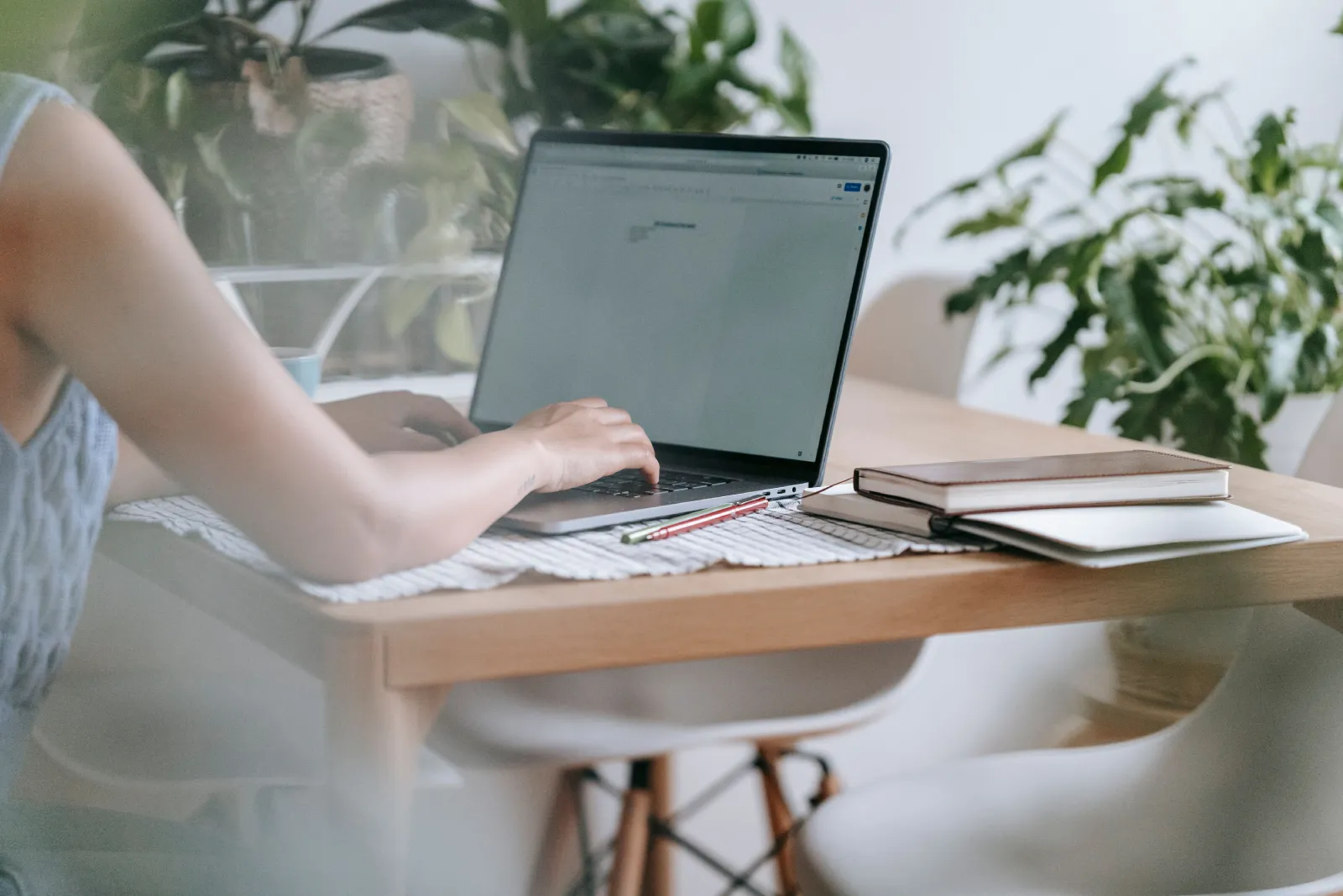 A woman typing on a laptop surrounded by plants and notebooks at her home workspace, creating a tranquil and focused environment