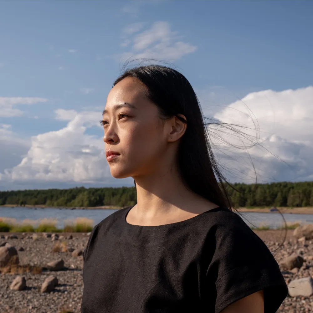 Portrait of an Asian woman in black shirt, looking afar by the rocky seaside under a cloudy sky.