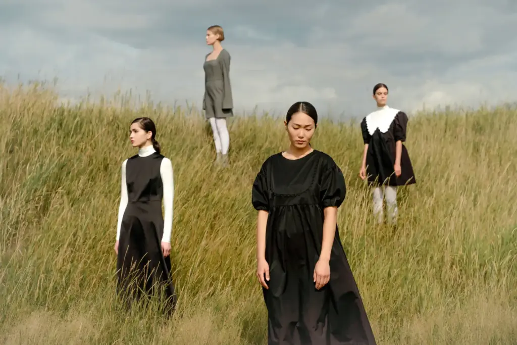 Four women in vintage-style dresses standing in a grassy field under a cloudy sky, outdoors