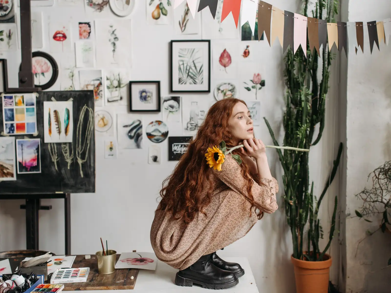 A redheaded woman in a floral dress sits in an art studio holding a sunflower.