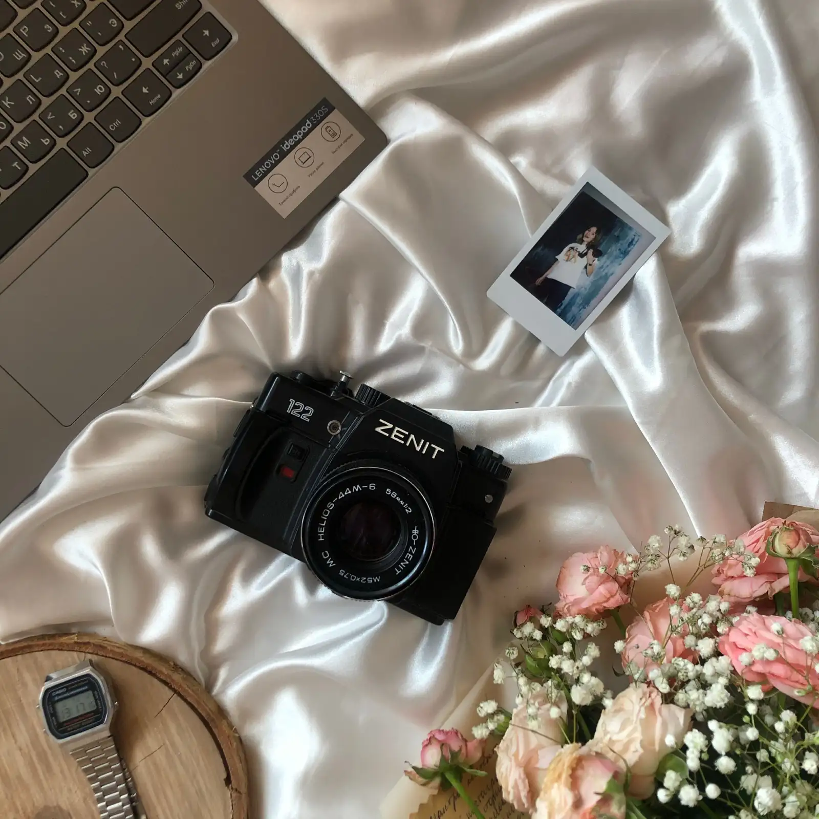 A vintage camera, flowers, and laptop on a silky white sheet, evoking romance