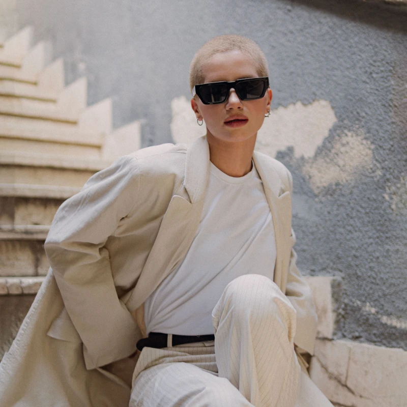 Chic woman in sunglasses and beige outfit sitting on stairs, modern urban fashion.