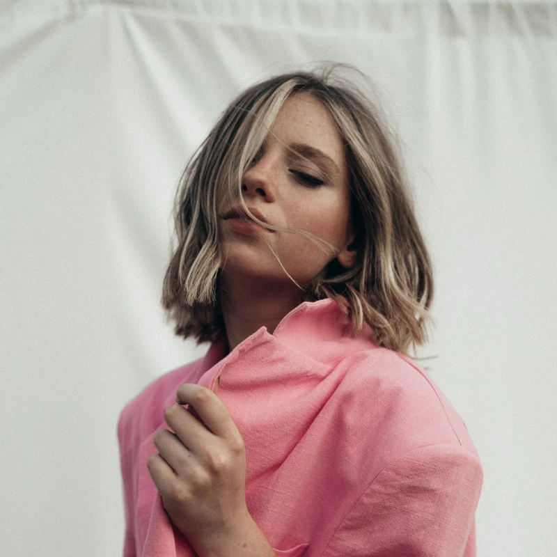 Young woman in a pink jacket outdoors, enjoying a serene moment with eyes closed.