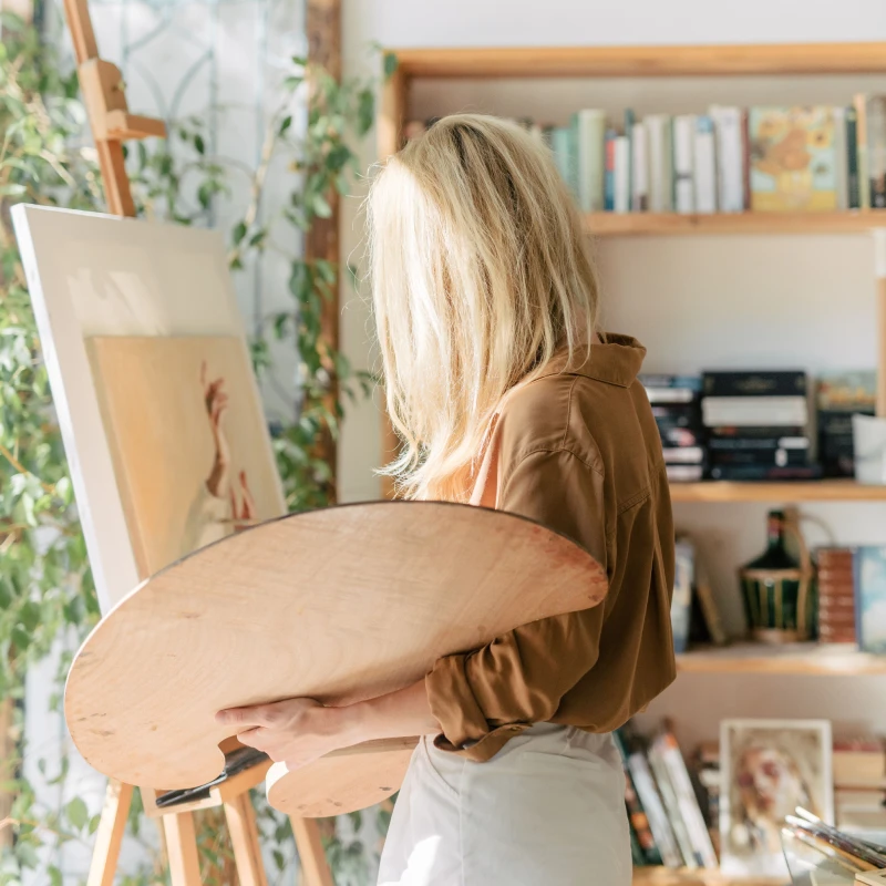 A woman artist with blonde hair paints on canvas in a cozy, sunlit studio filled with books and art supplies.